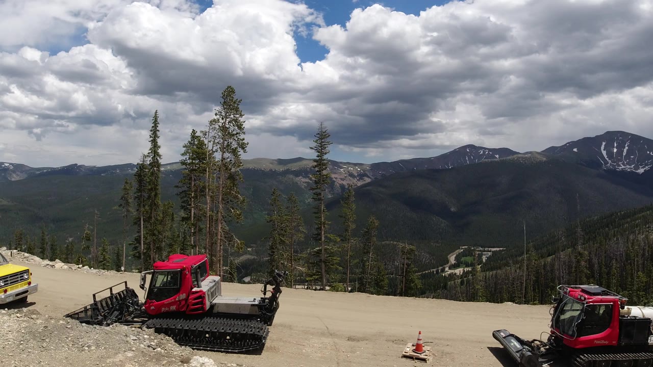 The second frame keeps the red tracked machine and yellow truck low in the view, with tall conifers, layered mountain ridges, and bright clouds over Winter Park, Colorado.
