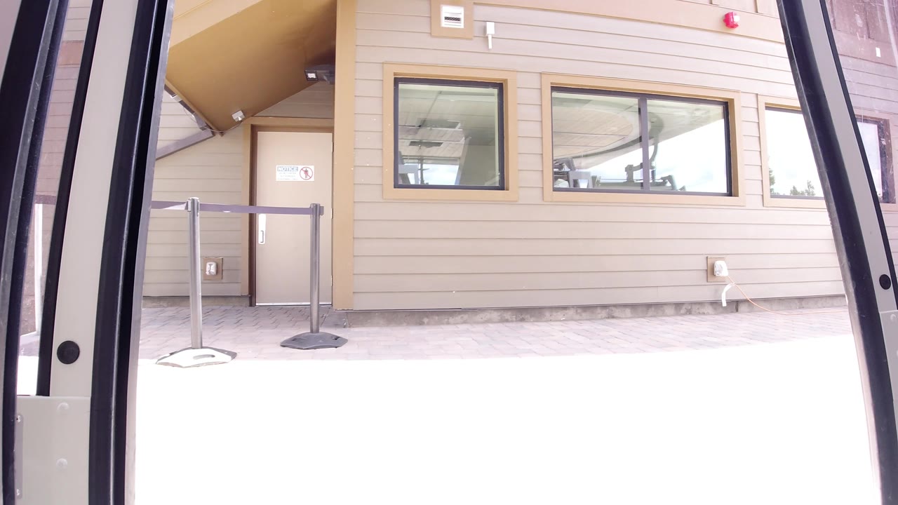 At 0:00 in Winter Park, Colorado, the view from inside a bus frames a yellow sided building with windows, a tan door, queue stanchions, bright foreground glare, and no visible people.