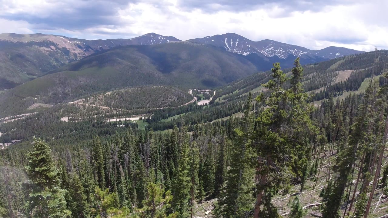 Winter Park, Colorado mountain valley seen over dense evergreens, with layered ridges, distant peaks carrying small snow patches, cloudy daylight, and no visible people.