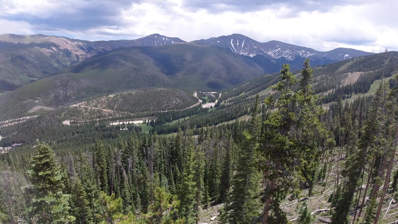 Winter Park, Colorado hillside view with conifer tops in the foreground, rolling forested ridges in the midground, a pale valley route, and no visible people.