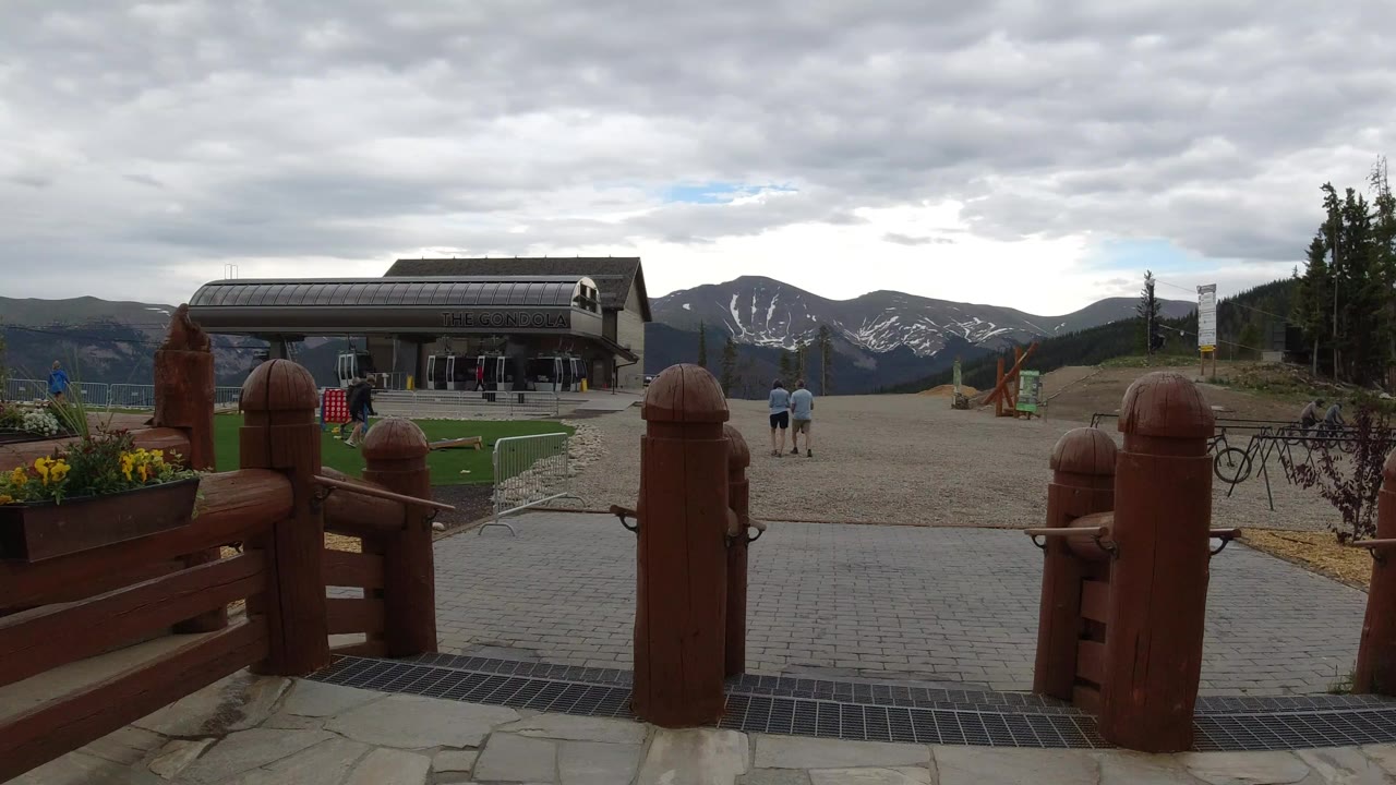 Winter Park, Colorado resort plaza seen past red-brown timber posts, with a base building, distant visitors, snow-streaked mountains, conifer slopes, and overcast sky.