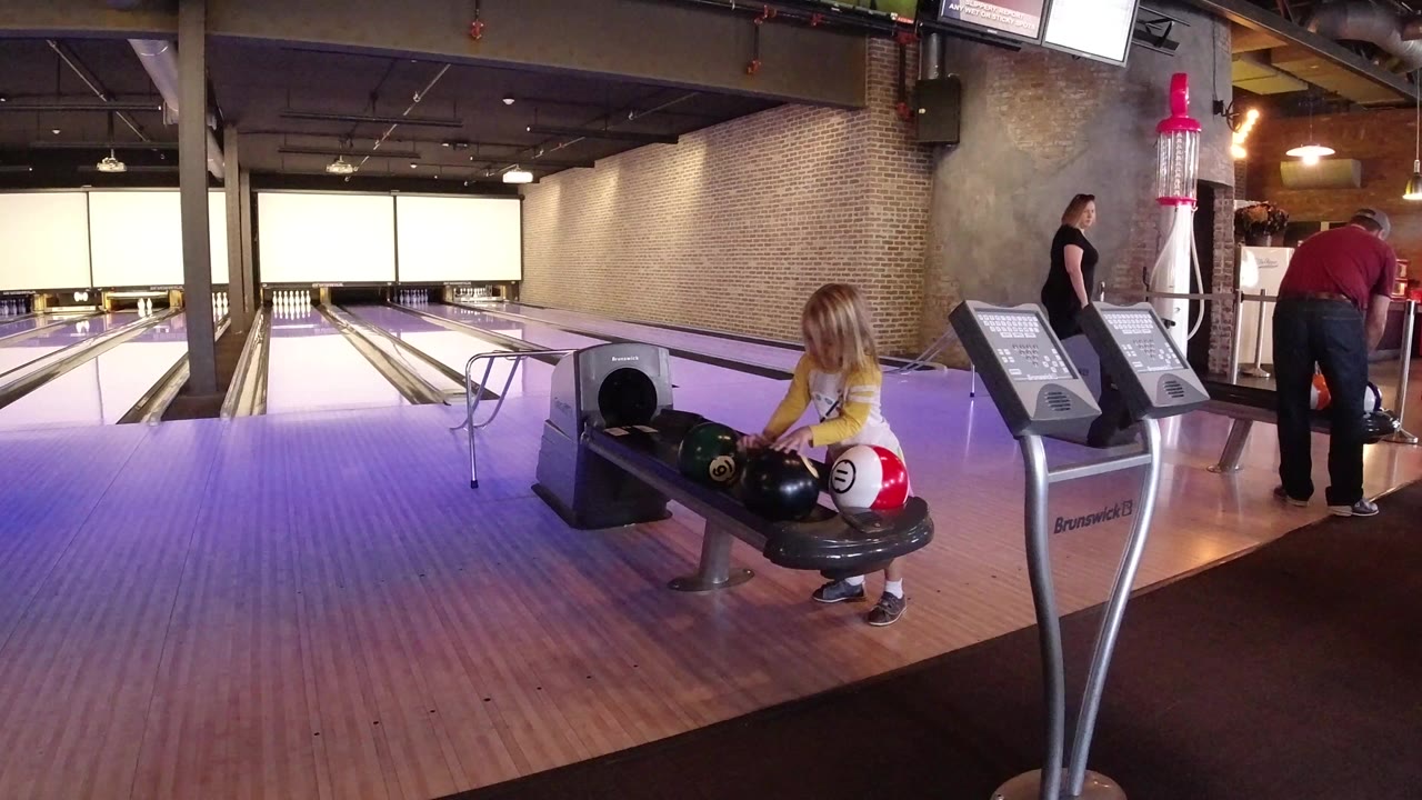 A young girl stands at a ball return in a Winter Park, Colorado bowling alley while adults and scoring consoles appear near the lanes behind her.