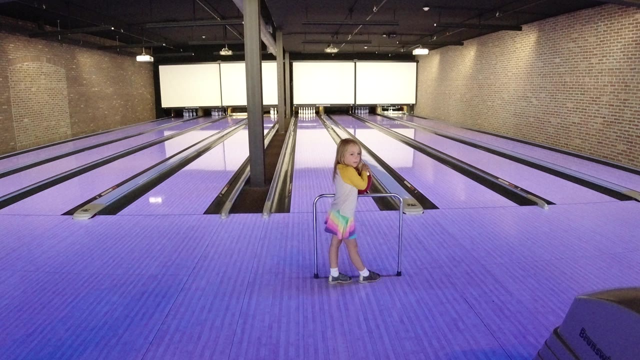 The child remains near the metal ramp in the Winter Park bowling alley, facing across the approach area toward the lanes and pin decks.