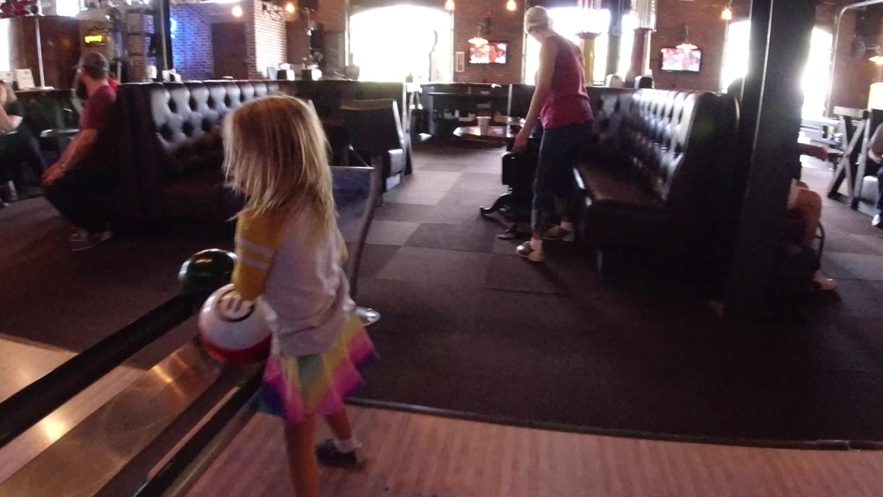 The young girl remains at the edge of the Winter Park bowling lane with a white ball in hand, while dark booth seating and other people occupy the background.
