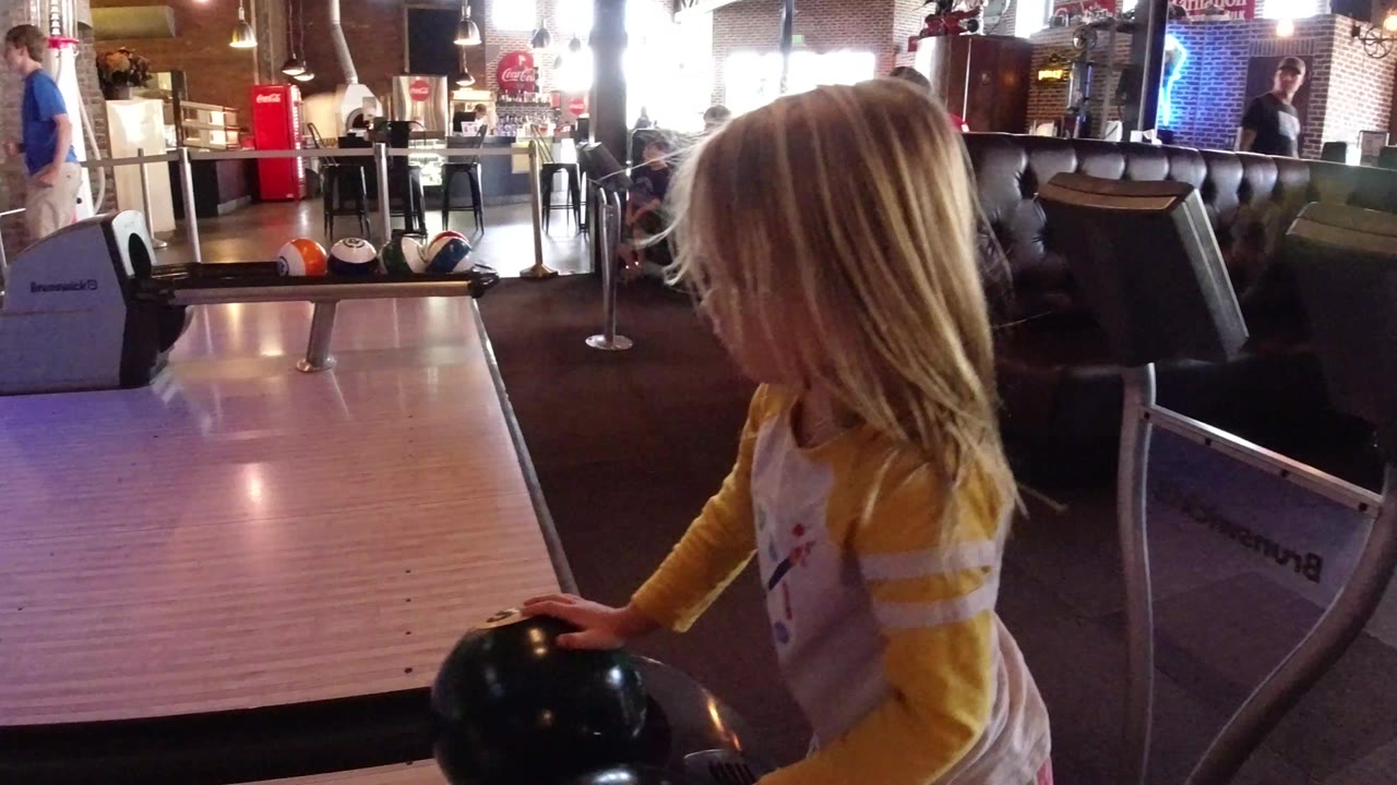 Young girl beside an indoor bowling lane holding a dark ball.