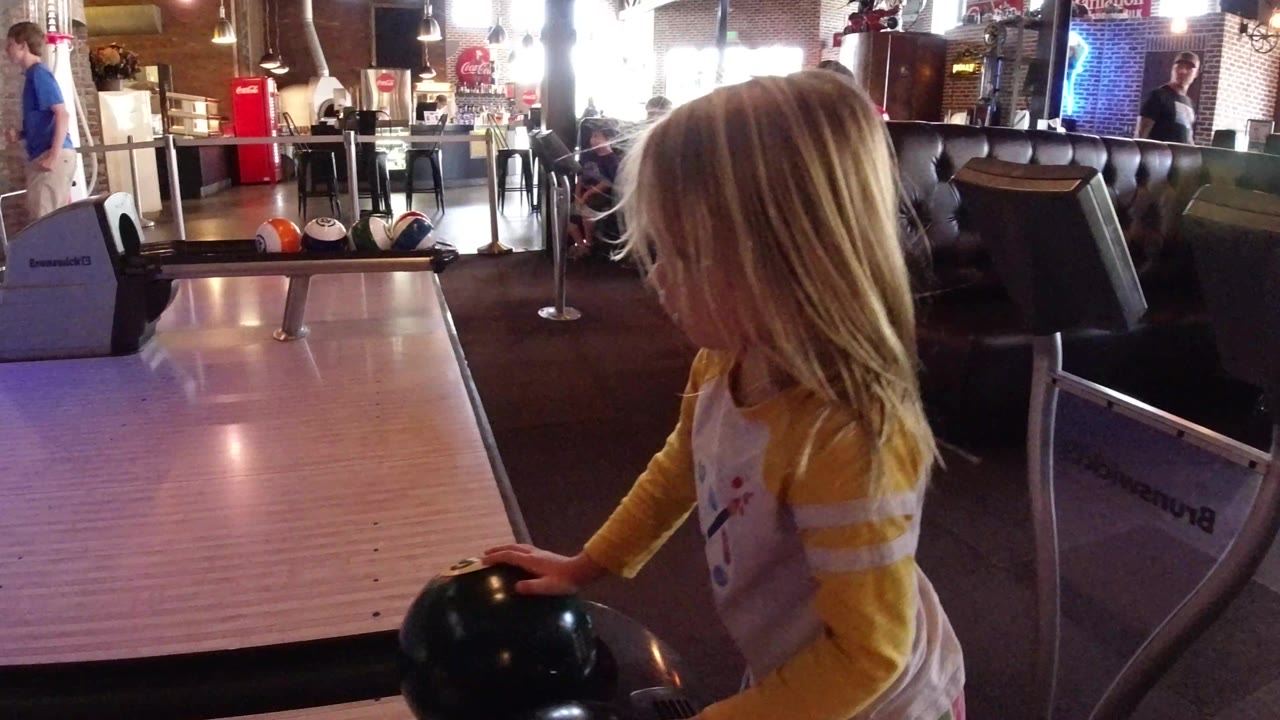 The girl remains near the lane edge in Winter Park, Colorado, facing the bowling surface while a dark ball rests under her hand and seating fills the background.