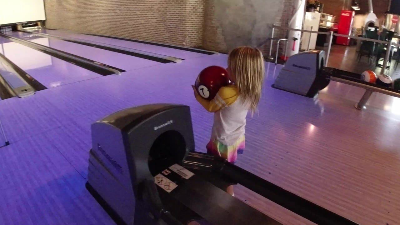 Blonde child holding a red bowling ball beside a ball return.