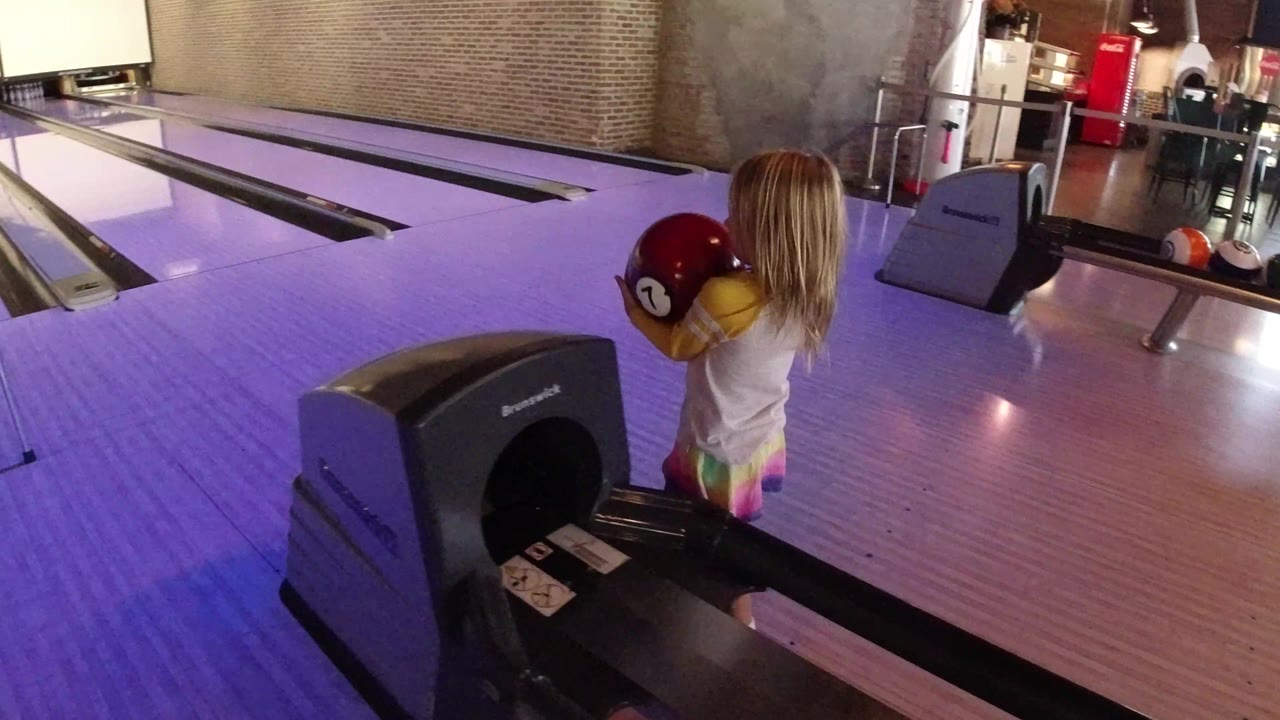 The child faces the bowling lanes with a red ball held at chest height, with the purple lane surface and ball return in the foreground.