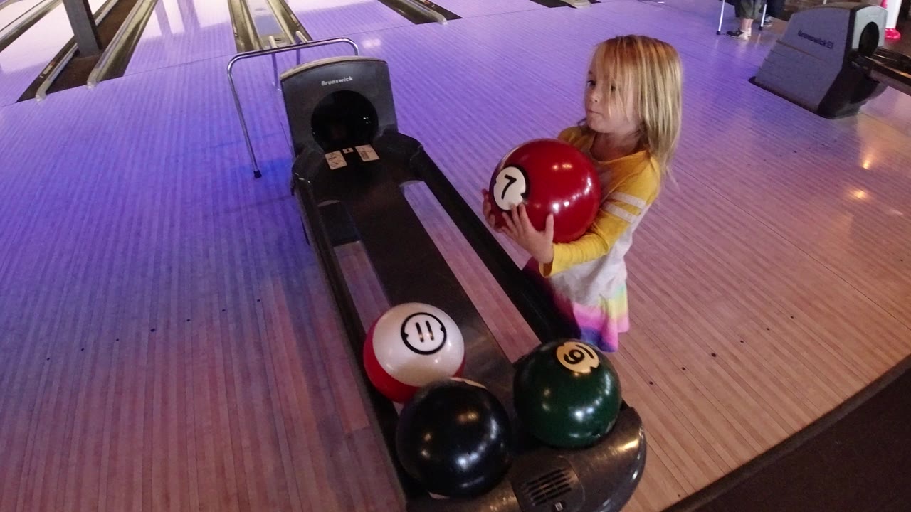 Child holding a red bowling ball beside a bowling lane return.
