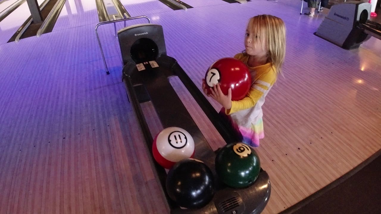 The child remains beside the bowling lane approach in Winter Park, Colorado, holding the red ball while black, white, and green balls sit on the return.