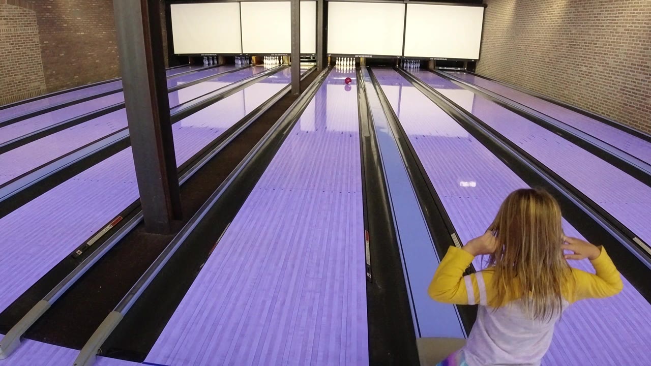 A young girl in Winter Park, Colorado faces the bowling pins with both hands lifted near her head, standing at the edge of a purple-lit lane.