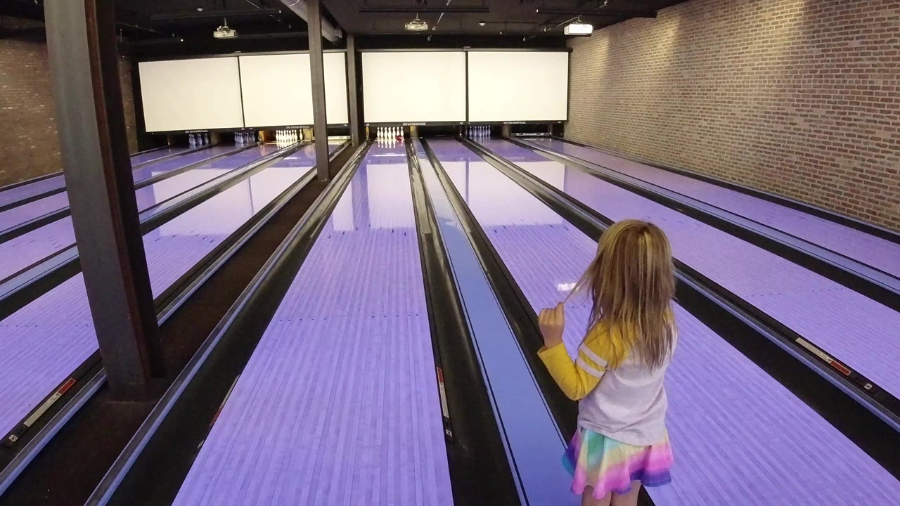 Child standing beside purple-lit bowling lanes in an indoor alley.