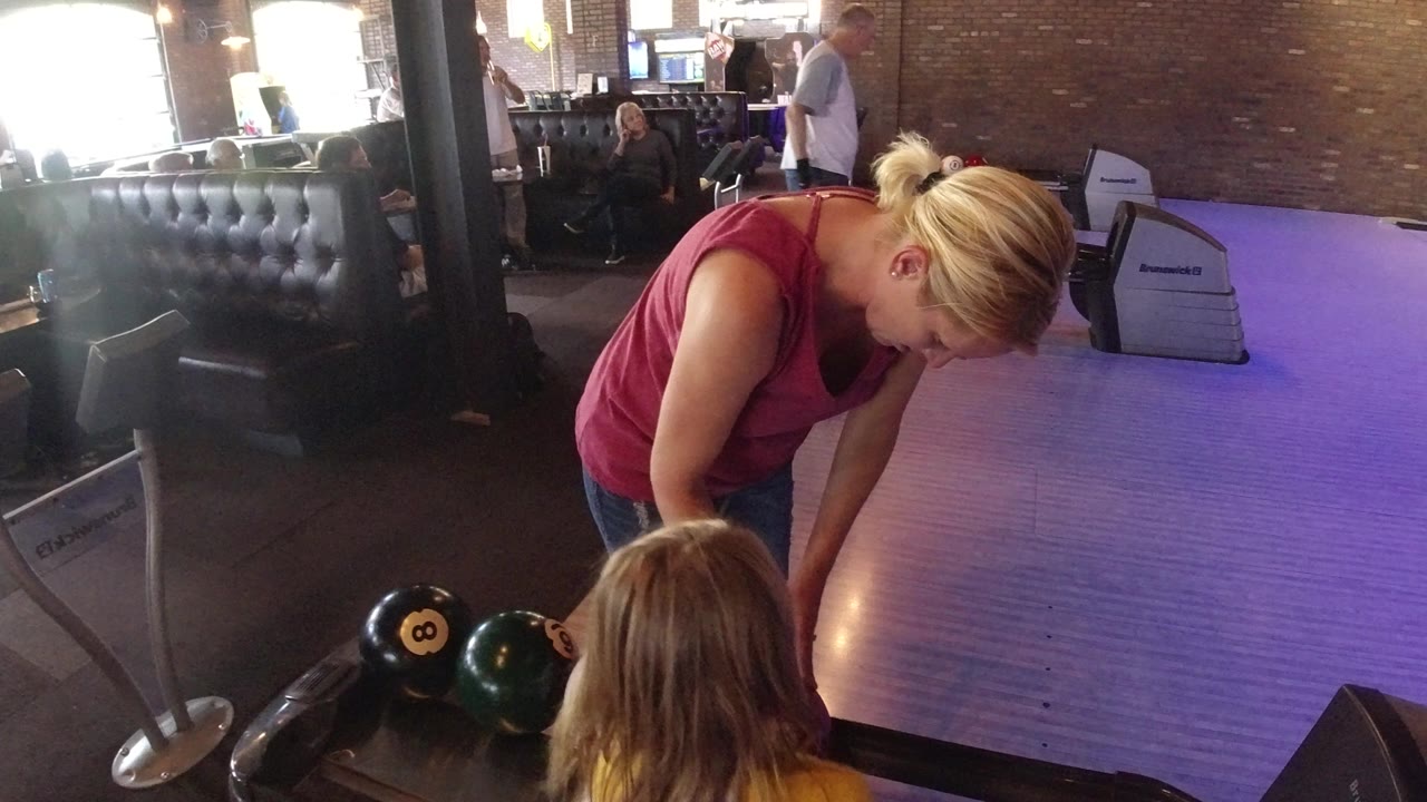 Woman leaning over bowling balls beside a purple-lit lane.