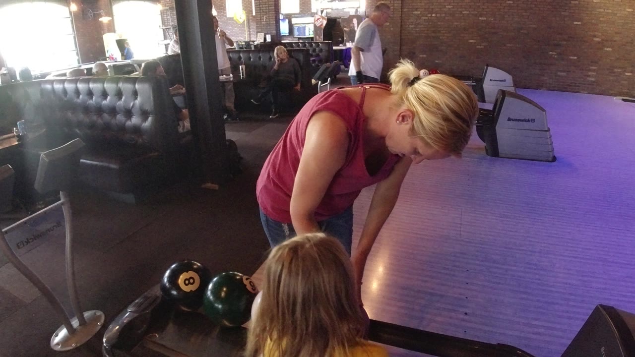 The woman remains bent over the ball return in the Winter Park bowling alley, with black, green, and red bowling balls visible near the lane edge.