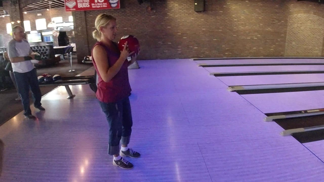 The woman keeps the red bowling ball near her chest while standing on the approach area, with multiple purple lanes and a brick wall visible in Winter Park, Colorado.