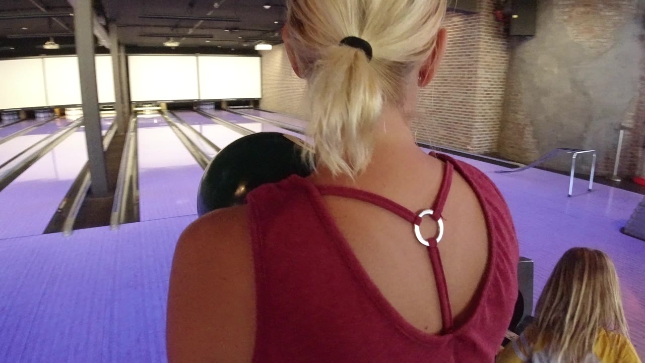 Blonde woman seen from behind holding a bowling ball near bowling lanes.