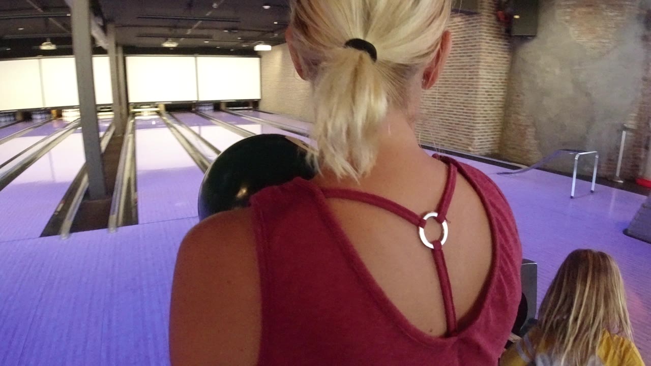 The woman remains framed from behind in the Winter Park bowling alley, with the ball tucked near her shoulder and a child low in the frame by the glowing lanes.