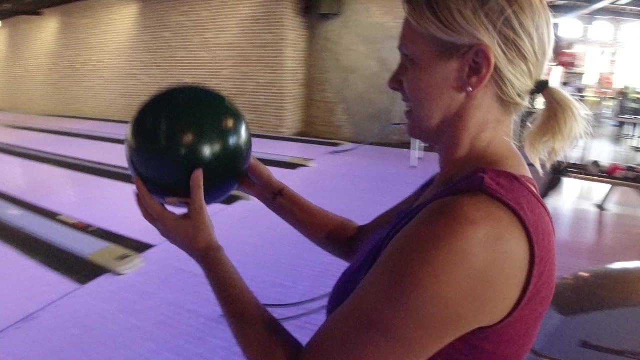The woman remains in side profile with the bowling ball raised, with the lane stretching behind her and warm lights reflecting off the purple lane surface.