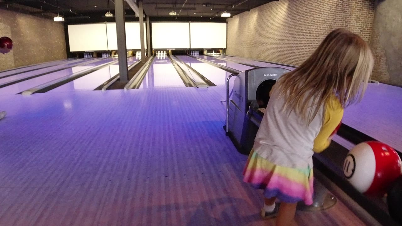 Young blond girl beside a bowling ball return at a purple-lit bowling alley.