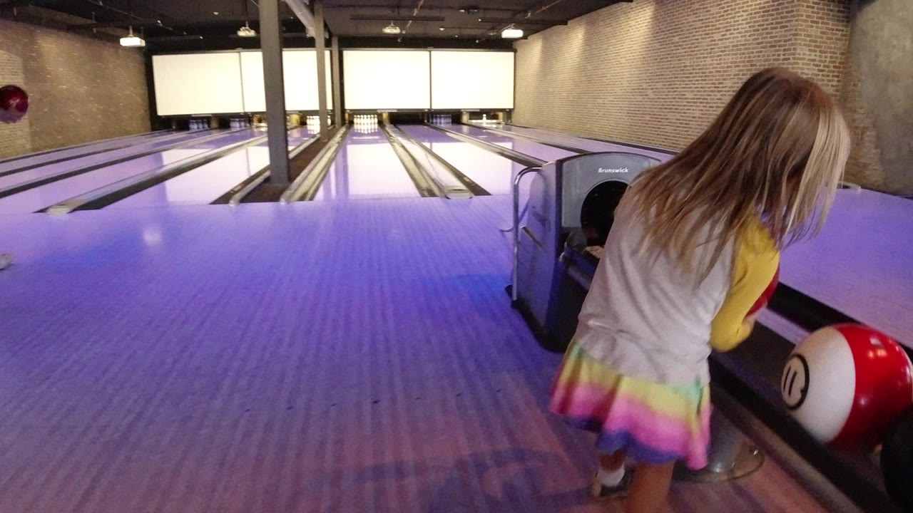 The girl leans beside the ball return at the bowling alley, with a red and white ball in the foreground and empty lanes visible ahead in Winter Park, Colorado.