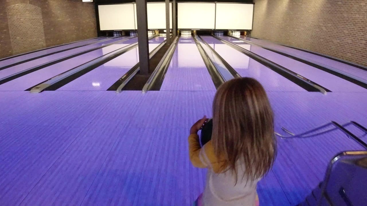 The child remains near the approach with a ball held at her side, looking toward the pins across the purple-lit bowling lanes in Winter Park, Colorado.