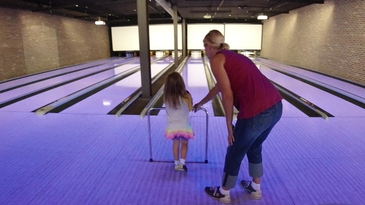 The child stands centered at the ramp while the adult leans in from the right, with several empty bowling lanes stretching toward white screens.