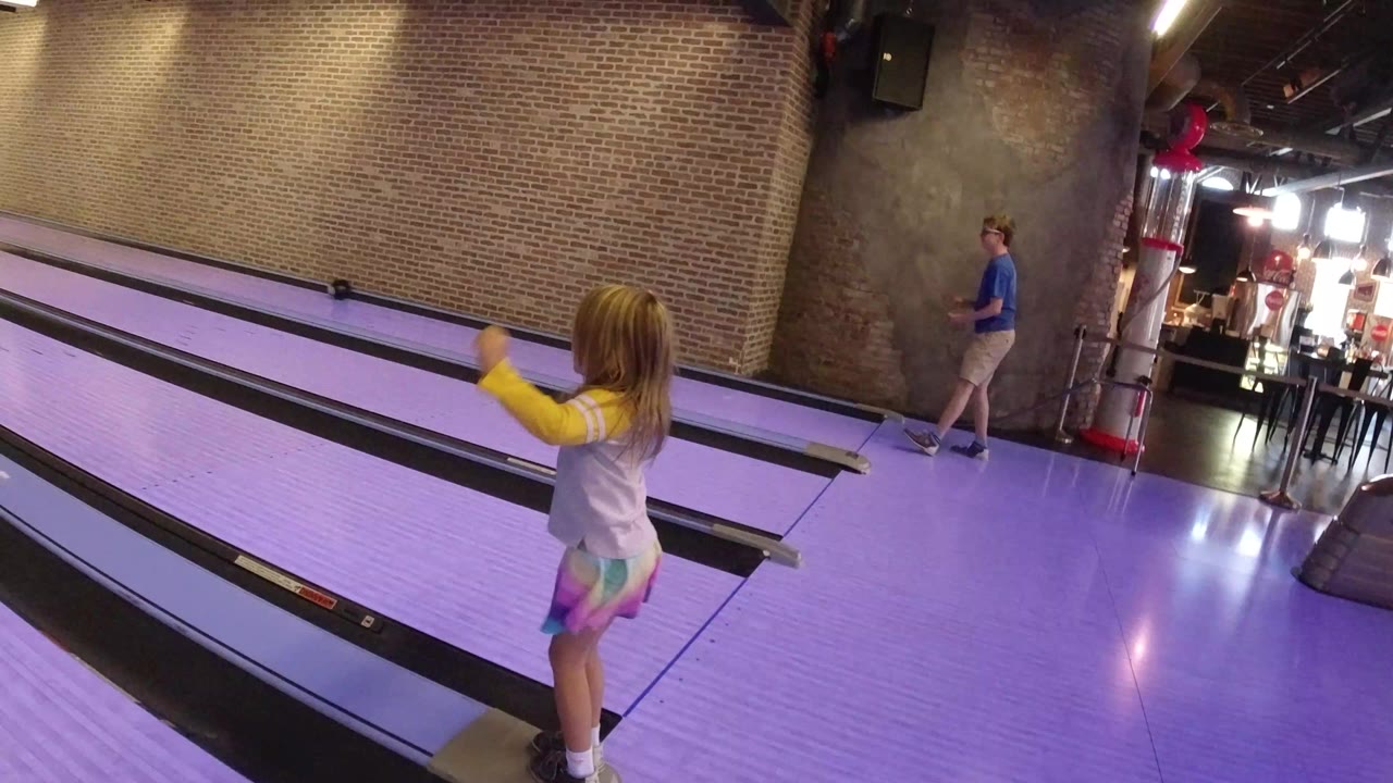 Young girl beside purple-lit bowling lanes with an adult in the background.