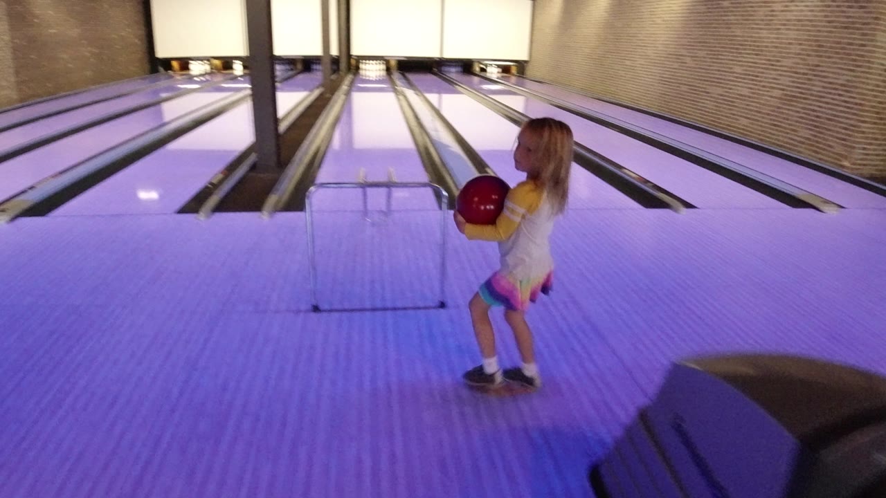 Small girl holding a red bowling ball beside purple-lit bowling lanes.