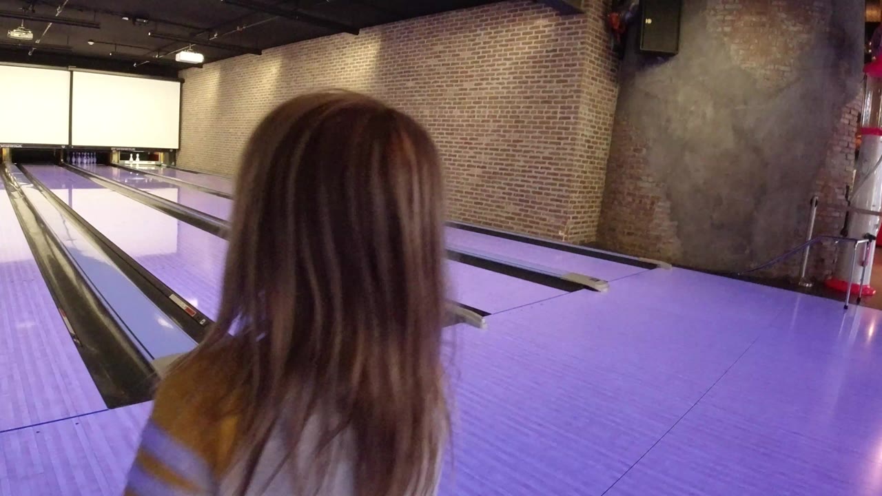 A young girl stands close to the bowling lane in Winter Park, Colorado, seen from behind with brick walls and bright scoring screens ahead.