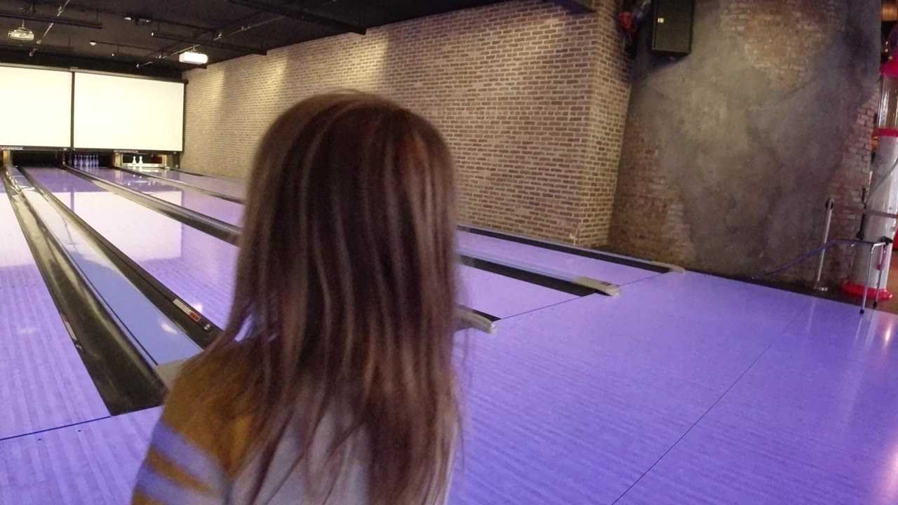 The girl remains in the foreground facing the lanes, with purple-lit boards, gutters, brick side walls, and pale scoring screens visible ahead in Winter Park, Colorado.