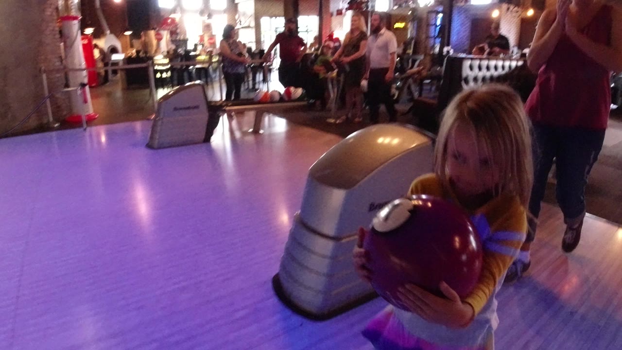 Young girl holding a purple bowling ball beside a glowing lane.