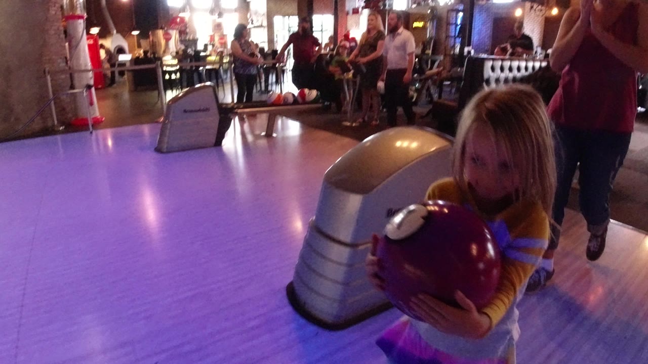 The young girl remains near the bowling lane approach in Winter Park, Colorado, holding the purple ball while adults stand and sit behind her under warm lights.