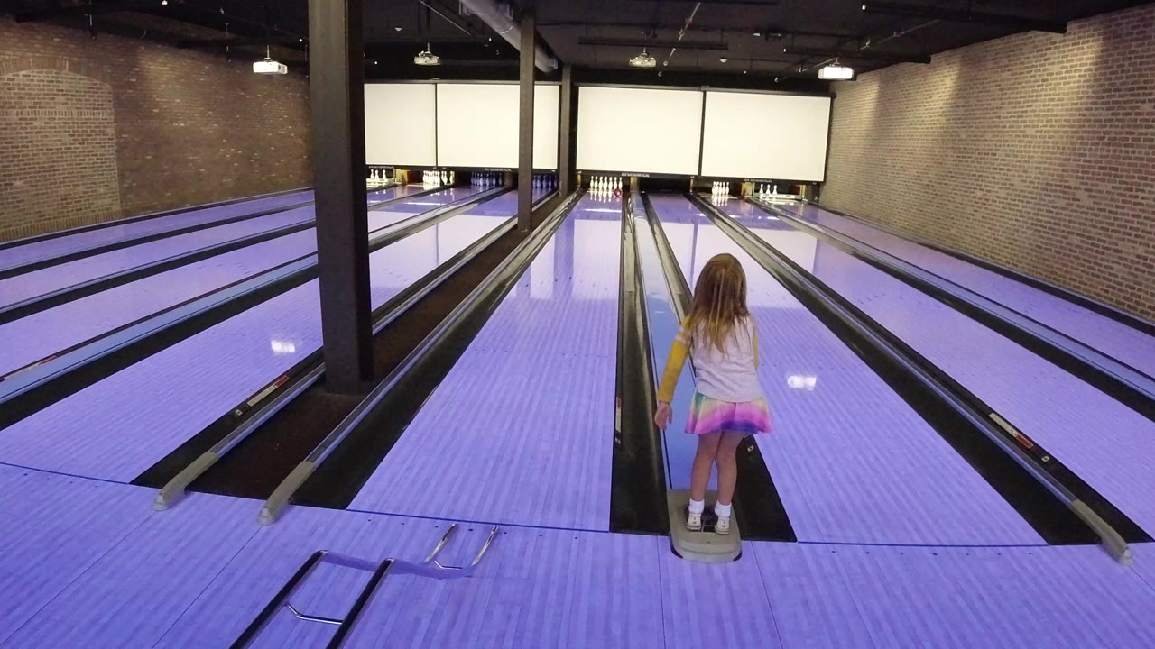 The girl stands centered between bowling lanes in Winter Park, Colorado, with purple-blue floor light and pins visible at the far end.