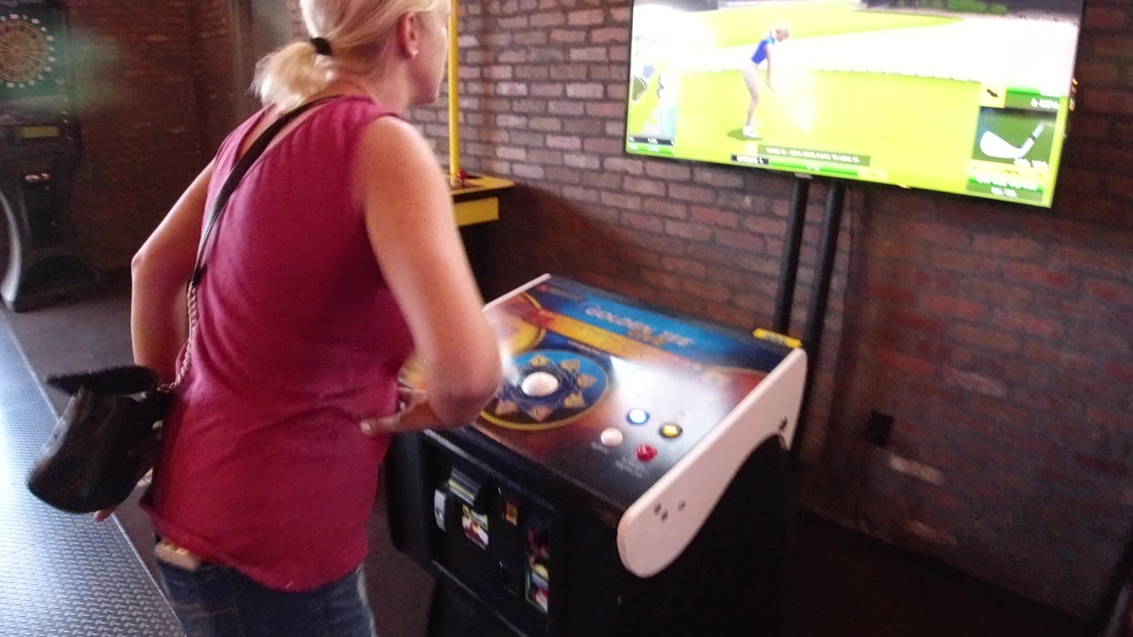 At 0:01 in Winter Park, Colorado, the woman faces the glowing golf game screen with her hands near the arcade cabinet controls and a brick wall behind the TV.