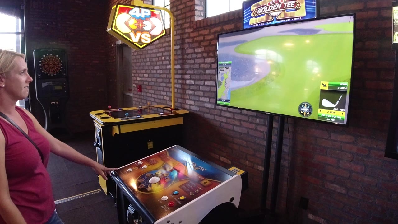 Blonde woman beside an arcade golf game screen in a brick-walled room.