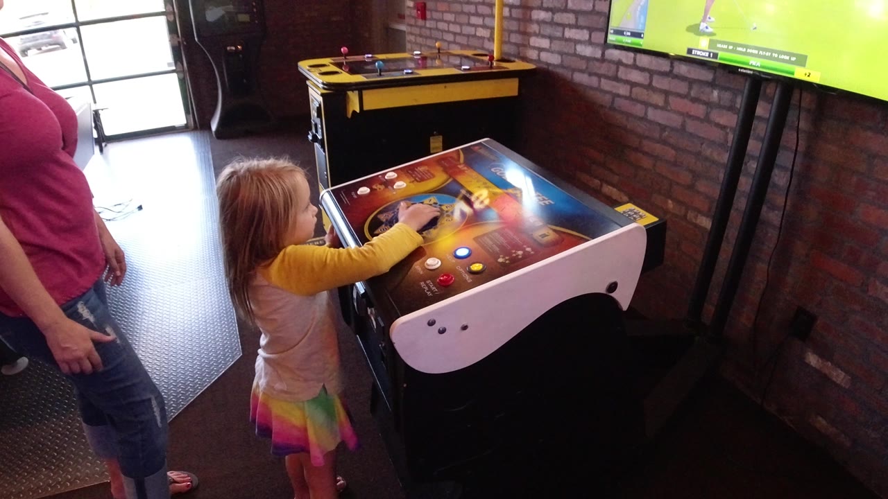 A child keeps one hand extended over the arcade controls as the adult stands beside the cabinet, with a sports game screen mounted on the brick wall in the background.