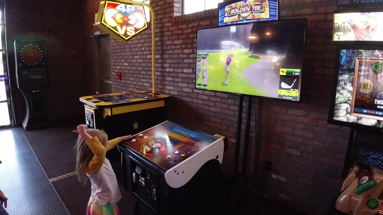 A young girl stands at an arcade control table in Winter Park, Colorado, watching a golf game screen under bright arcade signage in a brick-walled interior.