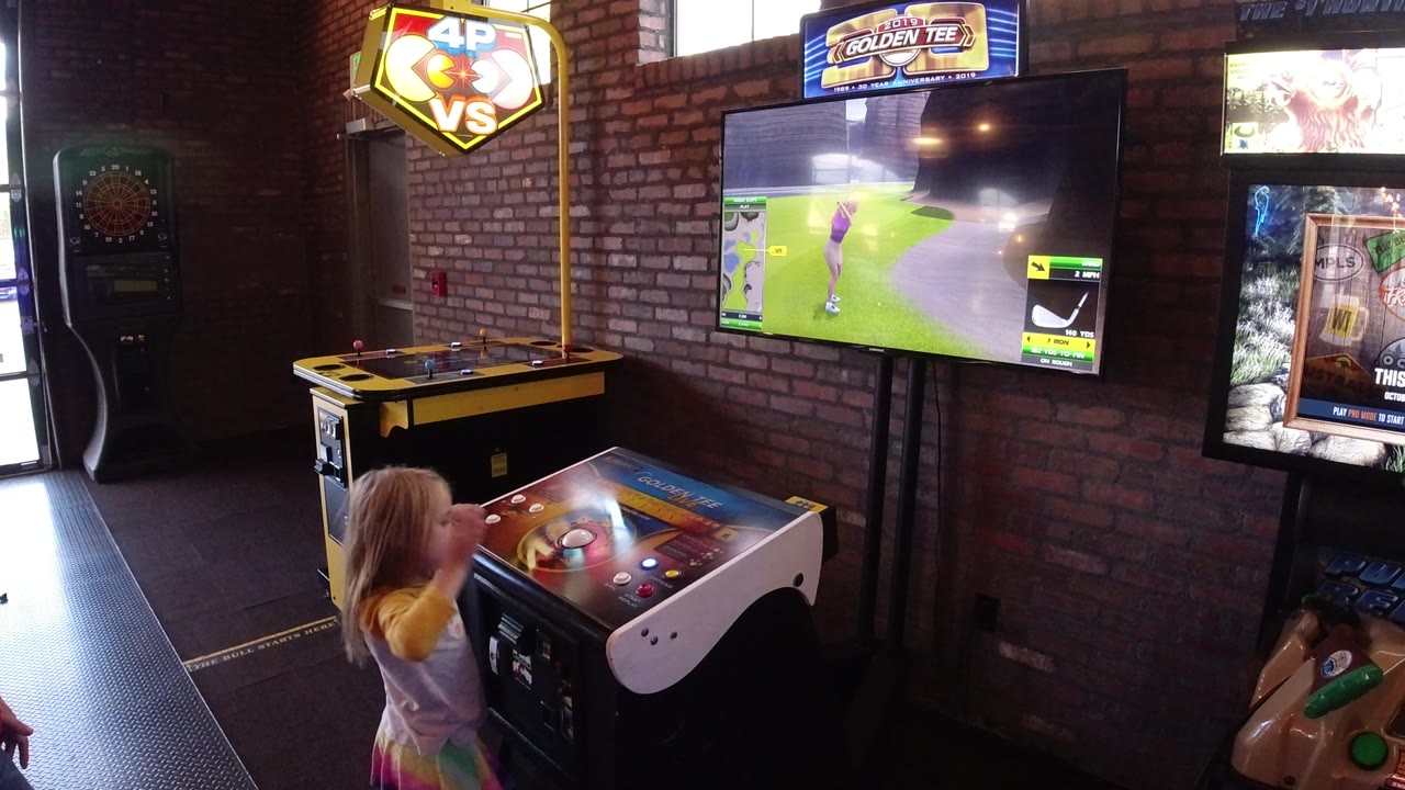The child stands at the right side of the arcade control deck in Winter Park, Colorado, with the golf game screen, brick wall, and lit arcade signs filling the room.