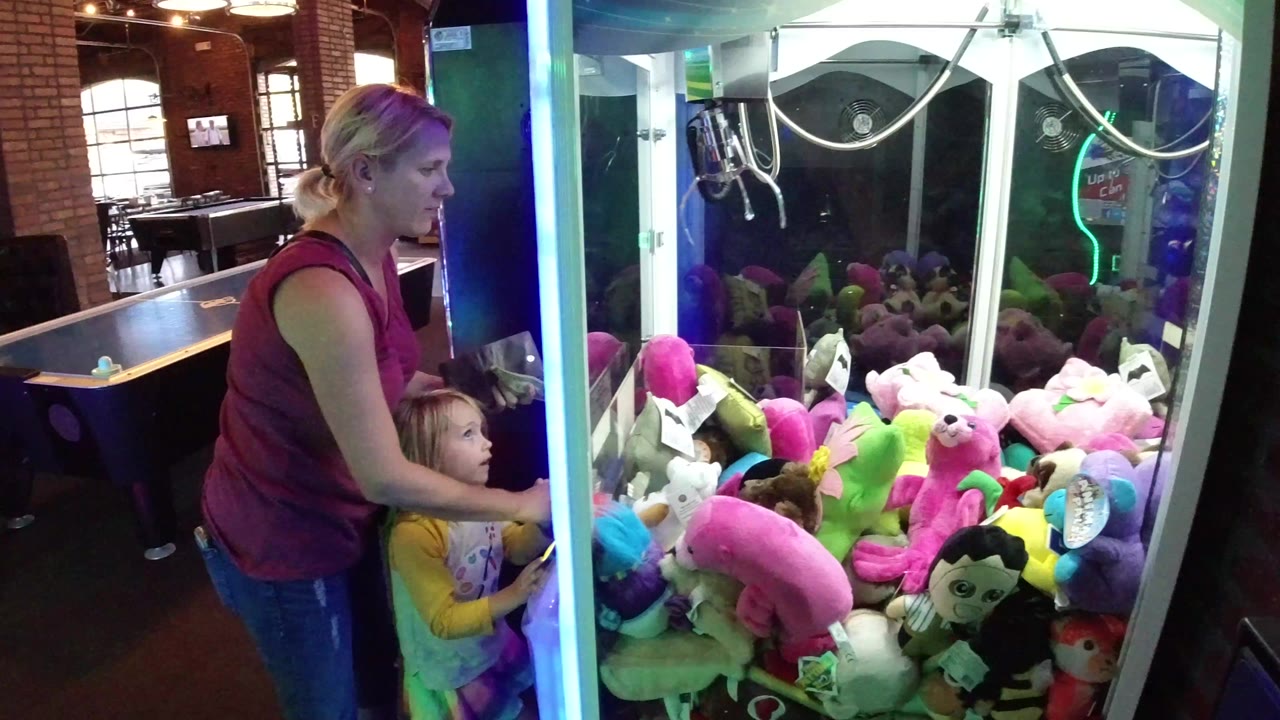 In Winter Park, Colorado, a mother and young daughter stand at the side of a glowing claw machine packed with pink, green, and white plush toys.