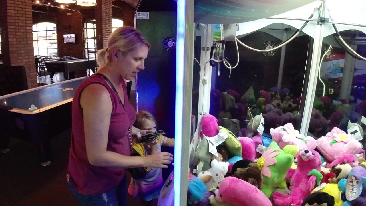 The woman looks down toward the young child at the controls while the claw machine glass reflects blue light and bright plush toys in Winter Park, Colorado.