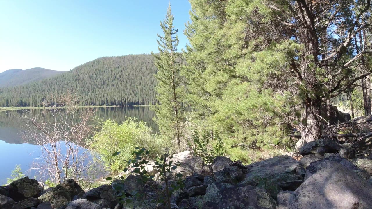 Winter Park, Colorado frame with boulders and low vegetation in front, dense evergreen branches to the right, still lake water to the left, and no visible people.