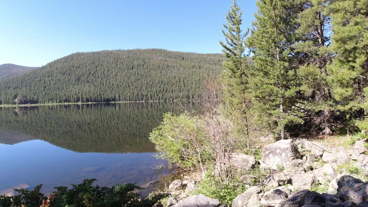 Wide Winter Park, Colorado lake view with a forested hillside reflected in still water, rocks and shrubs in the foreground, evergreens at right, and no visible people.