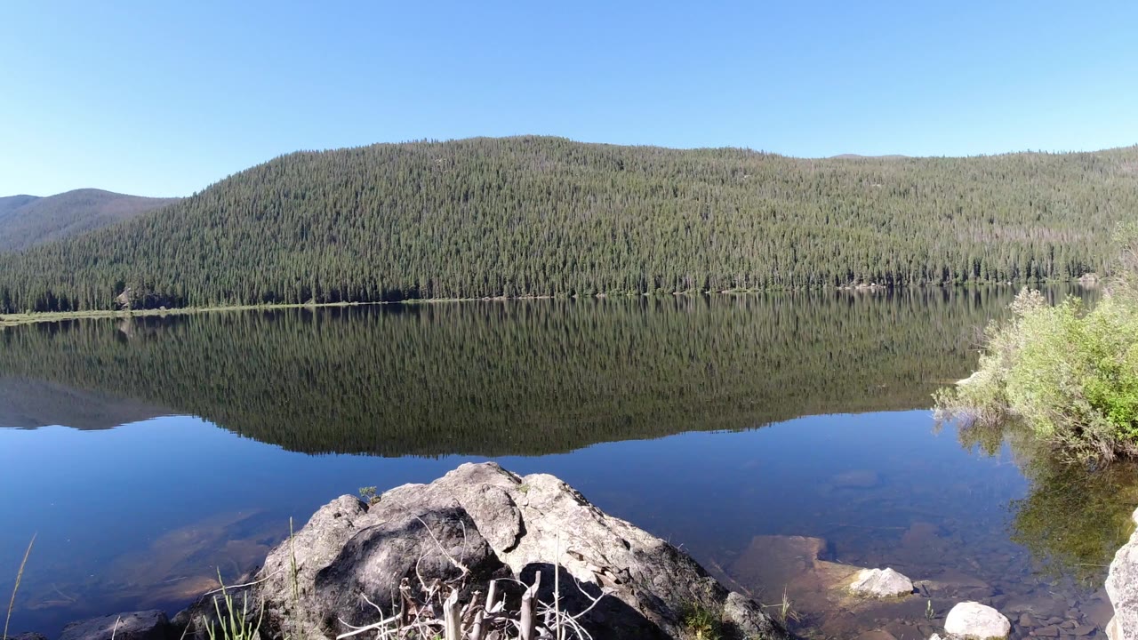 Winter Park, Colorado lake reflection with a forested hill across the water, blue sky overhead, shoreline rocks in the foreground, and no visible people.