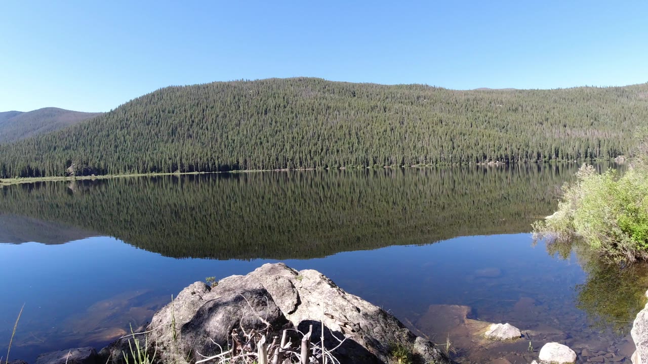 Winter Park, Colorado shoreline view with a broad forested hill reflected in smooth lake water, pale rocks at the bottom edge, and no visible people.