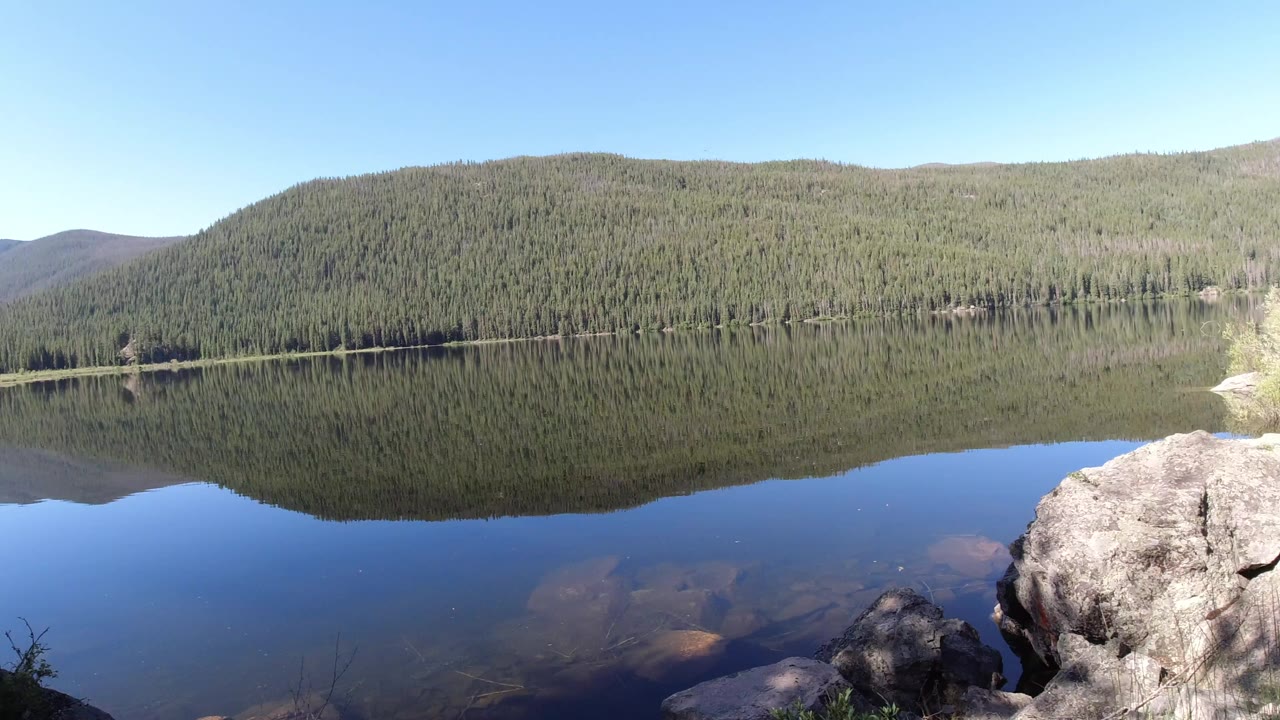 Wide Winter Park, Colorado lake view with evergreen hills mirrored in still water, foreground shoreline rocks at the lower right, and no visible people.