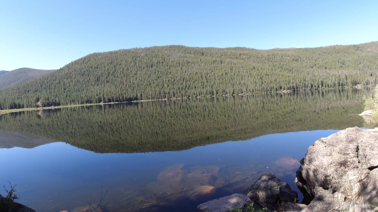 Winter Park, Colorado mountain lake with a broad tree-covered hill reflected across the water, pale shoreline at right, foreground boulders, and no visible people.