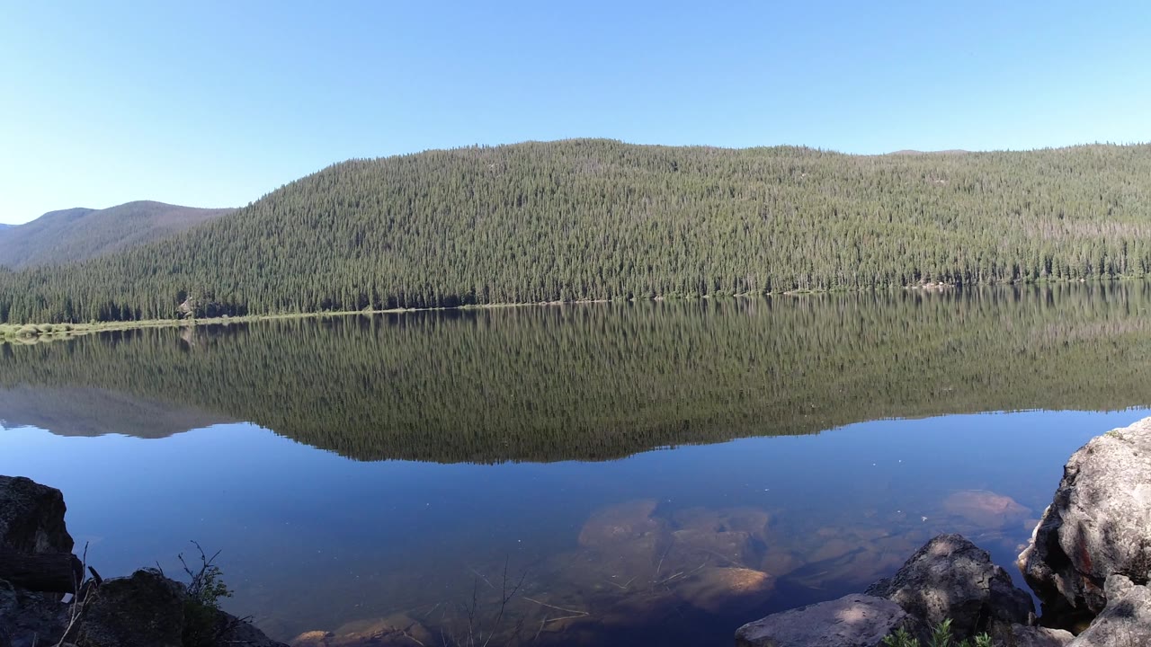 Lake and evergreen slope mirrored in clear water beside rocky foreground.