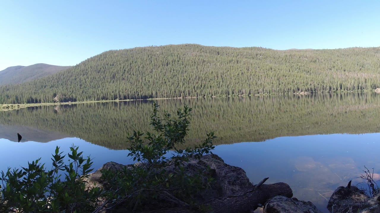 Wide Winter Park, Colorado lake view with a forested mountain ridge reflected in still water, foreground shrubs and rocks, and no visible people.