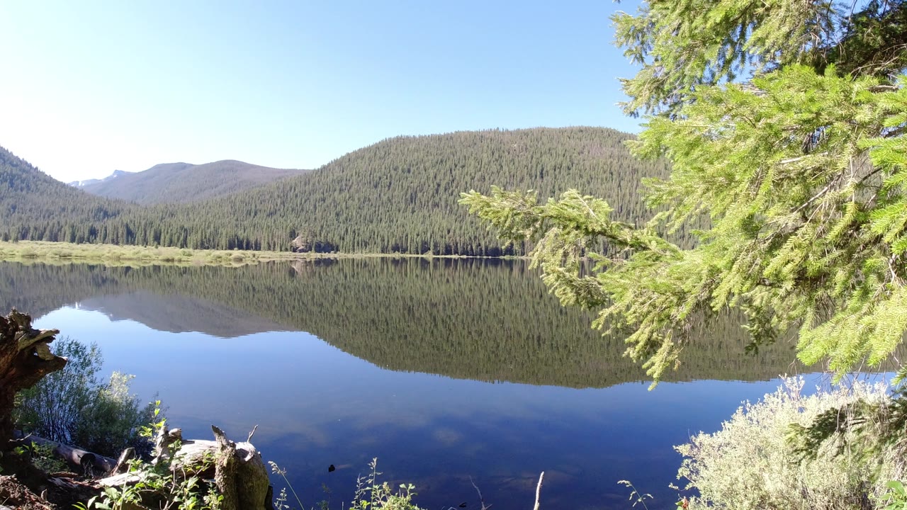 The lake remains still in Winter Park, Colorado, reflecting the tree-covered hills while an overhanging evergreen fills the right side of the frame and no visible people appear.