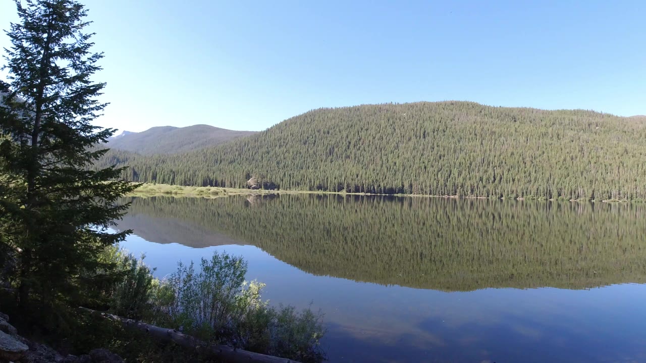 Mountain lake reflecting forested hills with evergreens at the left edge.